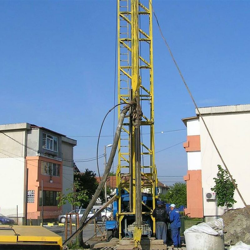 Turn de foraj galben între blocuri de locuințe, cu doi muncitori la bază și un furtun gros de noroi pe lângă turn; fotografie din arhiva firmei, localitatea nu e cunoscută